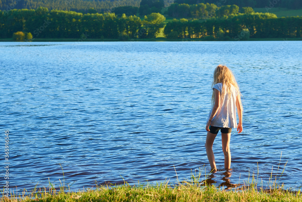 Φωτογραφία Alone child blond girl standing in the water in the light of the setting sun in
