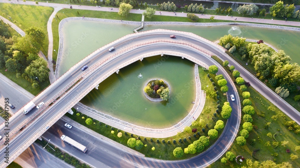 Aerial view of the roundabout on the river. Trees, overpass, buildings and vehicles can be seen. 