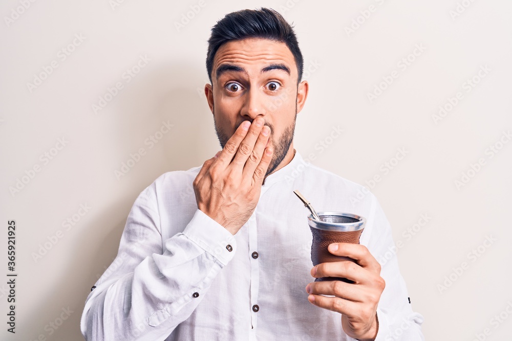 Young handsome man with beard drinking mate infusion beverage over white background covering mouth with hand, shocked and afraid for mistake. Surprised expression Young handsome man with beard drinking mate infusion beverage over white background covering mouth with hand, shocked and afraid for mistake. Surprised expression