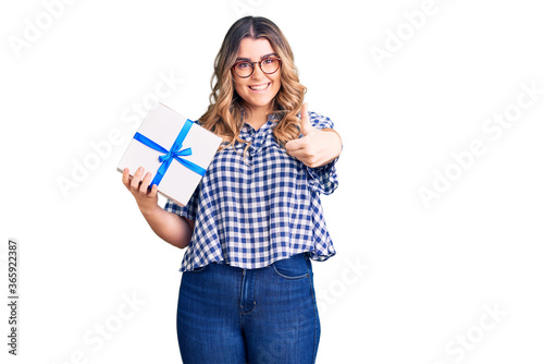 Young caucasian woman holding gift smiling happy and positive, thumb up doing excellent and approval sign