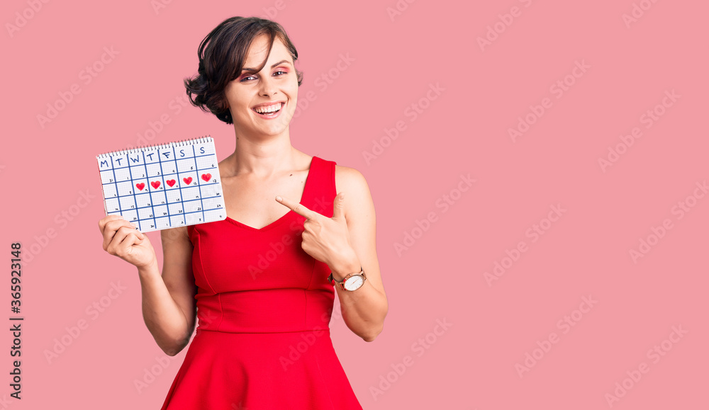 Beautiful young woman with short hair holding heart calendar smiling happy pointing with hand and finger Beautiful young woman with short hair holding heart calendar smiling happy pointing with hand and finger