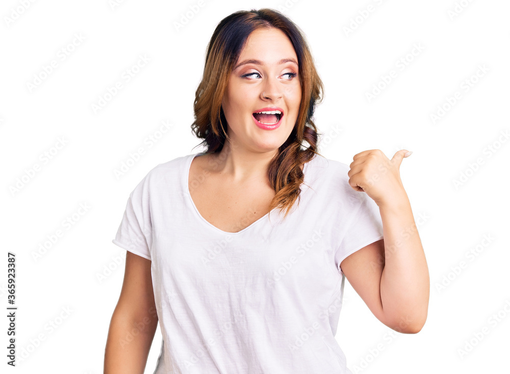 Young beautiful caucasian woman wearing casual white tshirt smiling with happy face looking and pointing to the side with thumb up. Young beautiful caucasian woman wearing casual white tshirt smiling with happy face looking and pointing to the side with thumb up.
