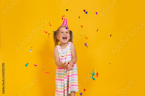 Happy smiling birthday child girl in pink cup surrounded by flying confetti on colored yellow background . Celebration, childhood, emotions.