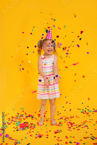 Happy smiling birthday child girl in pink cup surrounded by flying confetti on colored yellow background . Celebration, childhood, emotions.