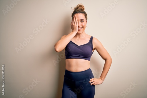 Young beautiful blonde sportswoman doing sport wearing sportswear over white background covering one eye with hand, confident smile on face and surprise emotion.