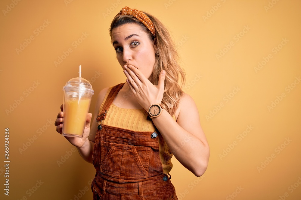 Young beautiful blonde woman drinking healthy glass of orange juice over yellow background cover mouth with hand shocked with shame for mistake, expression of fear, scared in silence, secret concept Young beautiful blonde woman drinking healthy glass of orange juice over yellow background cover mouth with hand shocked with shame for mistake, expression of fear, scared in silence, secret concept