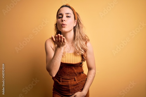 Young beautiful blonde woman wearing overalls and diadem standing over yellow background looking at the camera blowing a kiss with hand on air being lovely and sexy. Love expression.