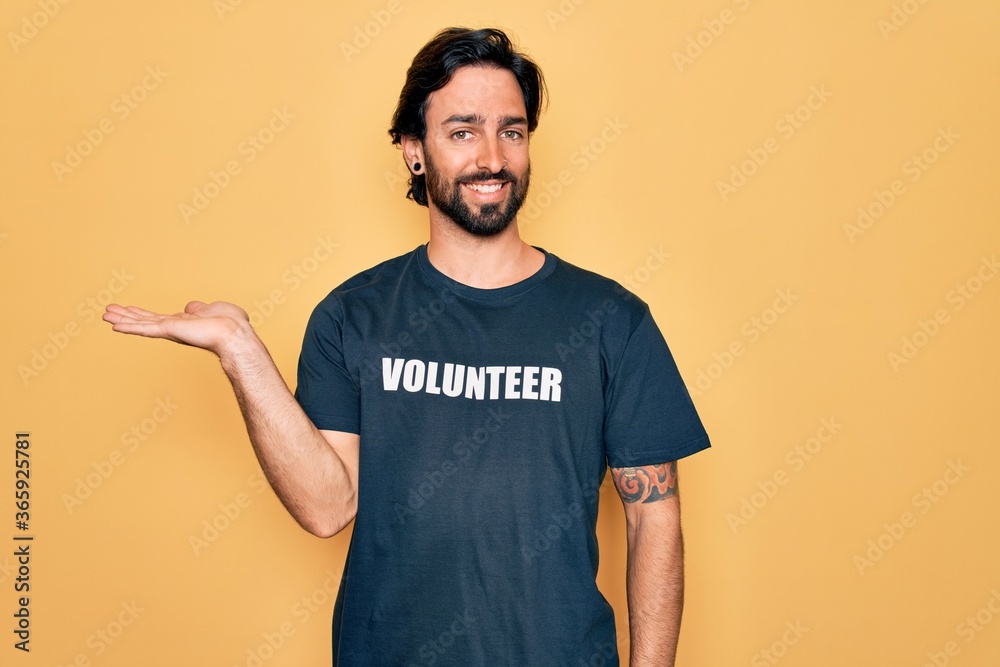 Young handsome hispanic volunteer man wearing volunteering t-shirt as social care smiling cheerful presenting and pointing with palm of hand looking at the camera. - obrazy, fototapety, plakaty 