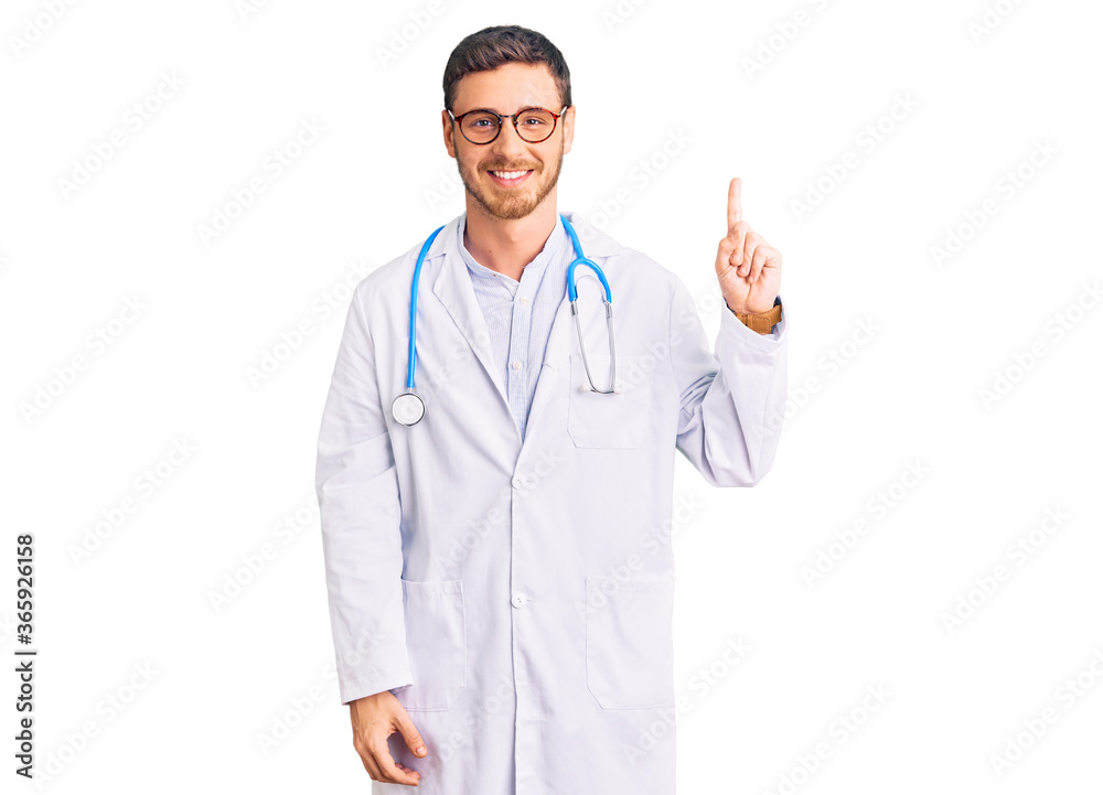 Handsome young man with bear wearing doctor uniform showing and pointing up with finger number one while smiling confident and happy.
