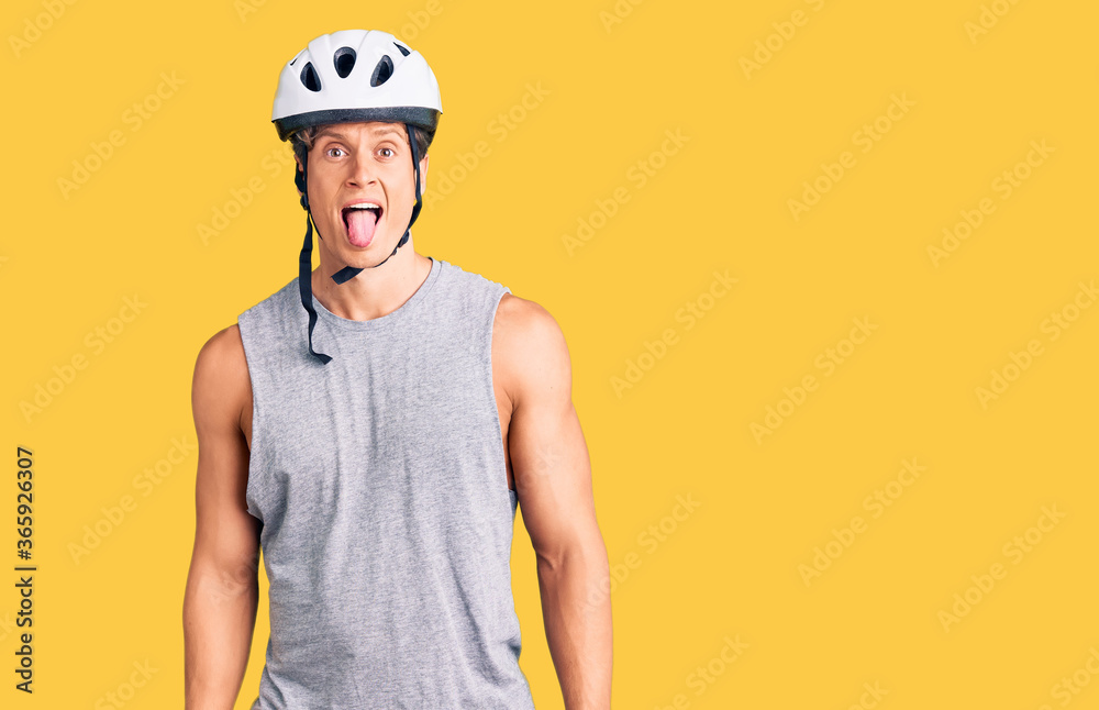 Young handsome man wearing bike helmet sticking tongue out happy with funny expression. emotion concept.