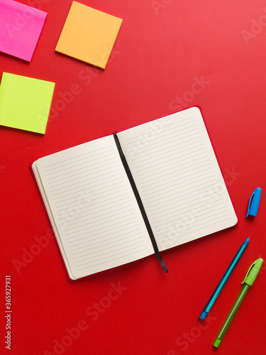 Top view of an open blank red notebook in the center, colored reminders in high corner and blue and green pen in the bottom corner, in a red background, with space for text