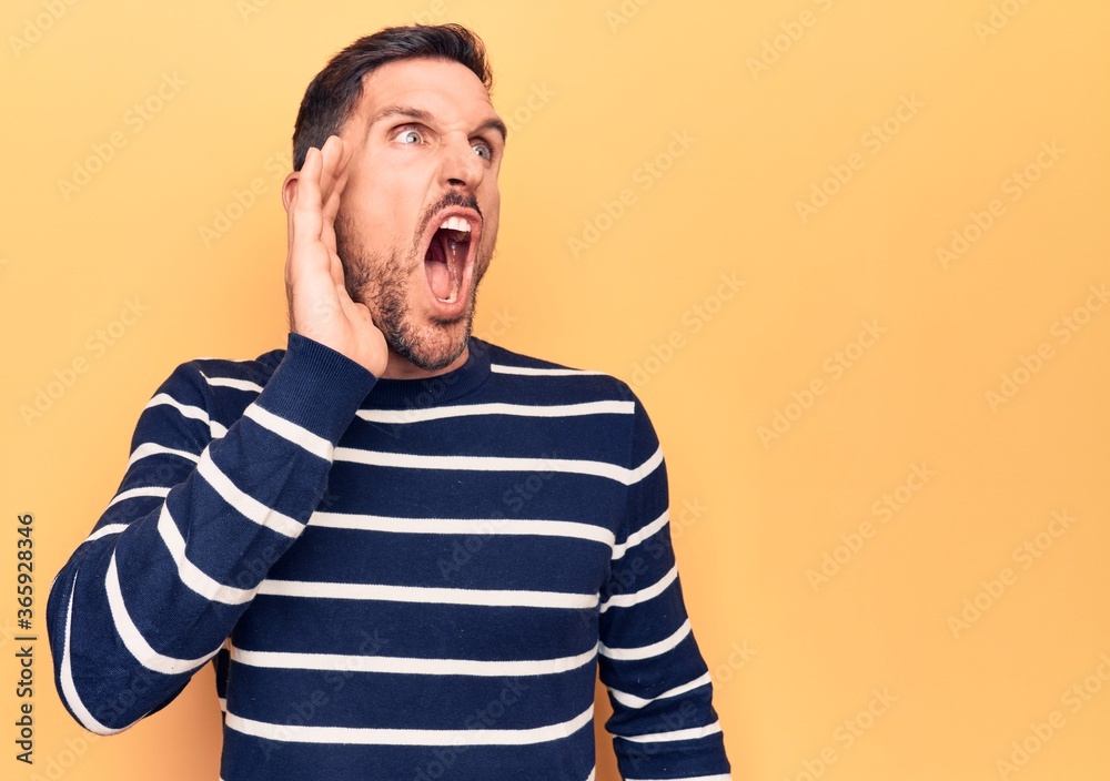 Obraz premium Young handsome man wearing casual striped sweater standing over isolated yellow background shouting and screaming loud to side with hand on mouth. Communication concept.