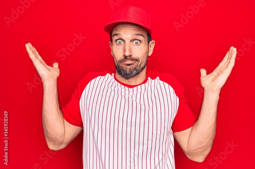 Young handsome player man wearing baseball sportswear over isolated red background clueless and confused with open arms, no idea and doubtful face.