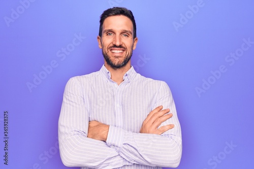 Young handsome man wearing casual striped shirt standing over isolated purple background happy face smiling with crossed arms looking at the camera. Positive person.