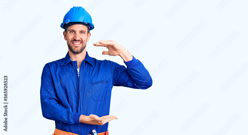Young handsome man wearing worker uniform and hardhat gesturing with hands showing big and large size sign, measure symbol. smiling looking at the camera. measuring concept.
