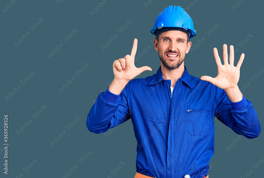 Young handsome man wearing worker uniform and hardhat showing and pointing up with fingers number seven while smiling confident and happy.
