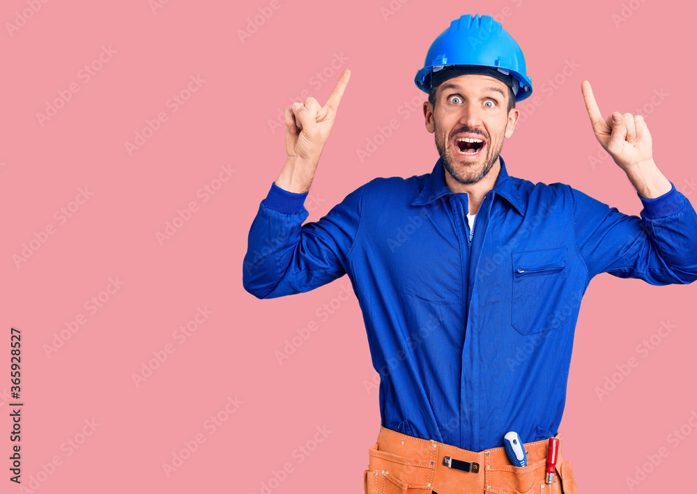 Young handsome man wearing worker uniform and hardhat smiling amazed and surprised and pointing up with fingers and raised arms.
