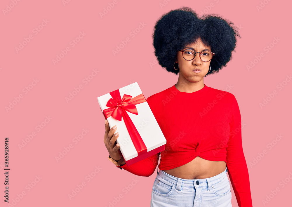 Young african american girl holding gift puffing cheeks with funny face. mouth inflated with air, crazy expression. Young african american girl holding gift puffing cheeks with funny face. mouth inflated with air, crazy expression.