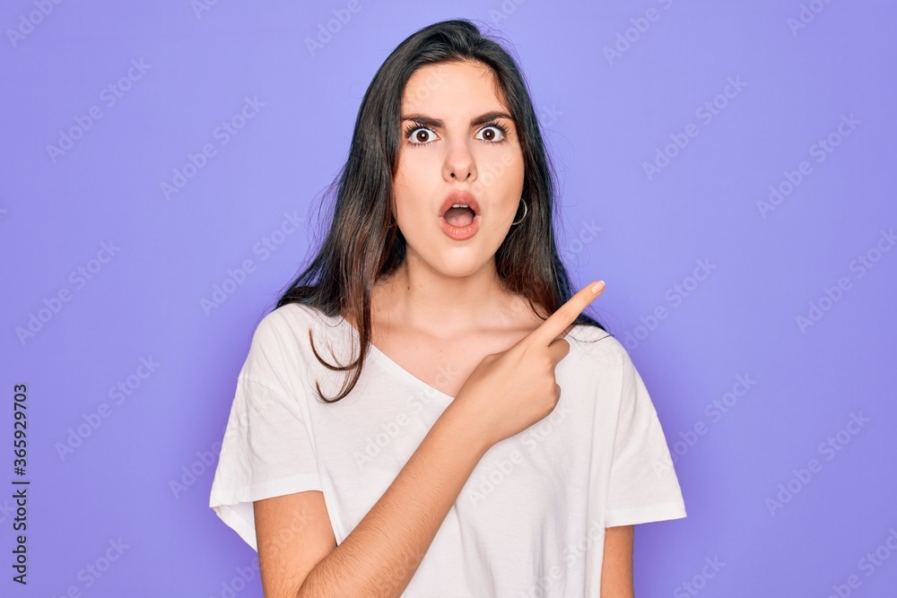 Young beautiful brunette woman wearing casual white t-shirt over purple background Surprised pointing with finger to the side, open mouth amazed expression. Young beautiful brunette woman wearing casual white t-shirt over purple background Surprised pointing with finger to the side, open mouth amazed expression.