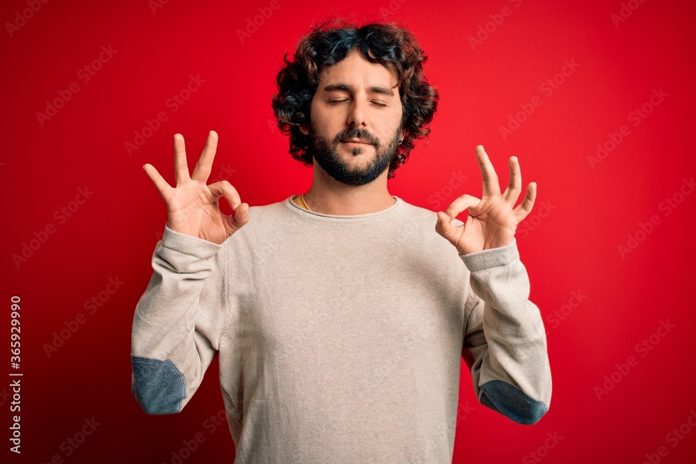 Naklejka premium Young handsome man with beard wearing casual sweater standing over red background relax and smiling with eyes closed doing meditation gesture with fingers. Yoga concept.