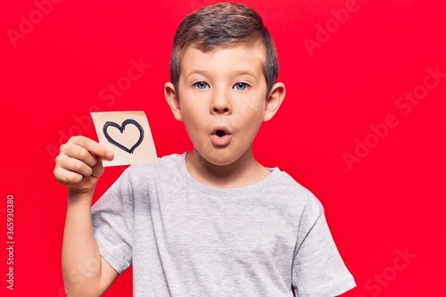 Cute blond kid holding heart reminder scared and amazed with open mouth for surprise, disbelief face