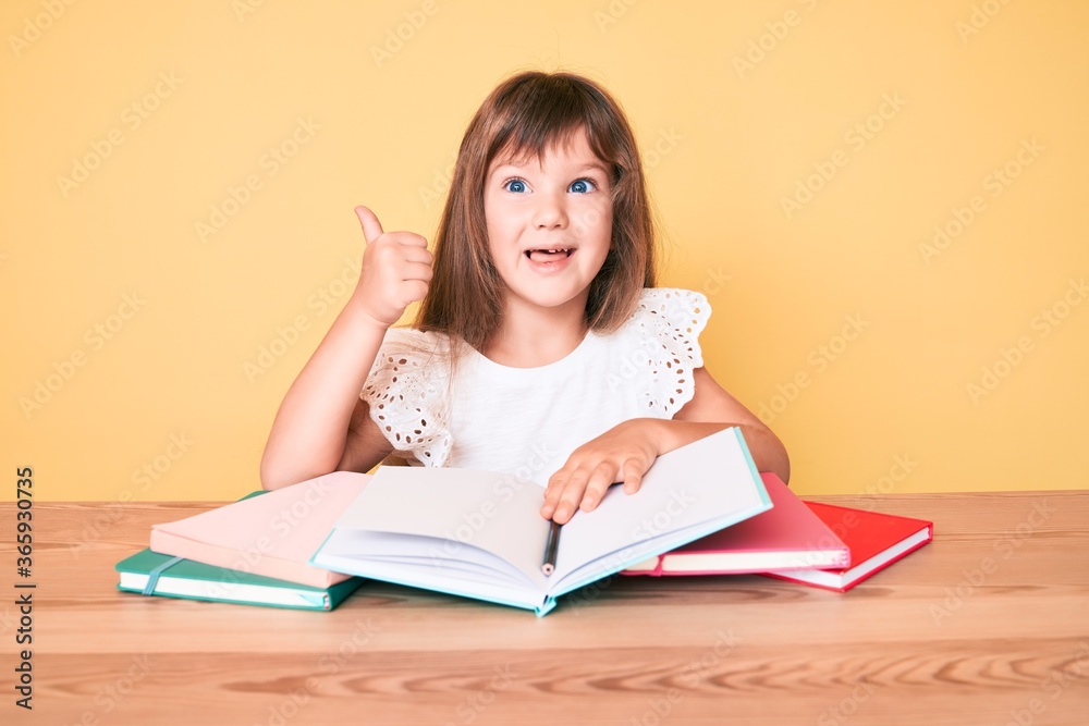 Little caucasian kid girl with long hair studying for school exam smiling happy and positive, thumb up doing excellent and approval sign Little caucasian kid girl with long hair studying for school exam smiling happy and positive, thumb up doing excellent and approval sign