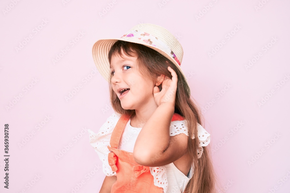 Little caucasian kid girl with long hair wearing spring hat smiling with hand over ear listening and hearing to rumor or gossip. deafness concept.
