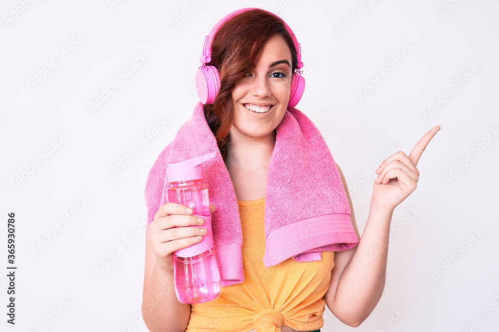 Young beautiful woman wearing towel and headphones holding bottle of water smiling happy pointing with hand and finger to the side Young beautiful woman wearing towel and headphones holding bottle of water smiling happy pointing with hand and finger to the side