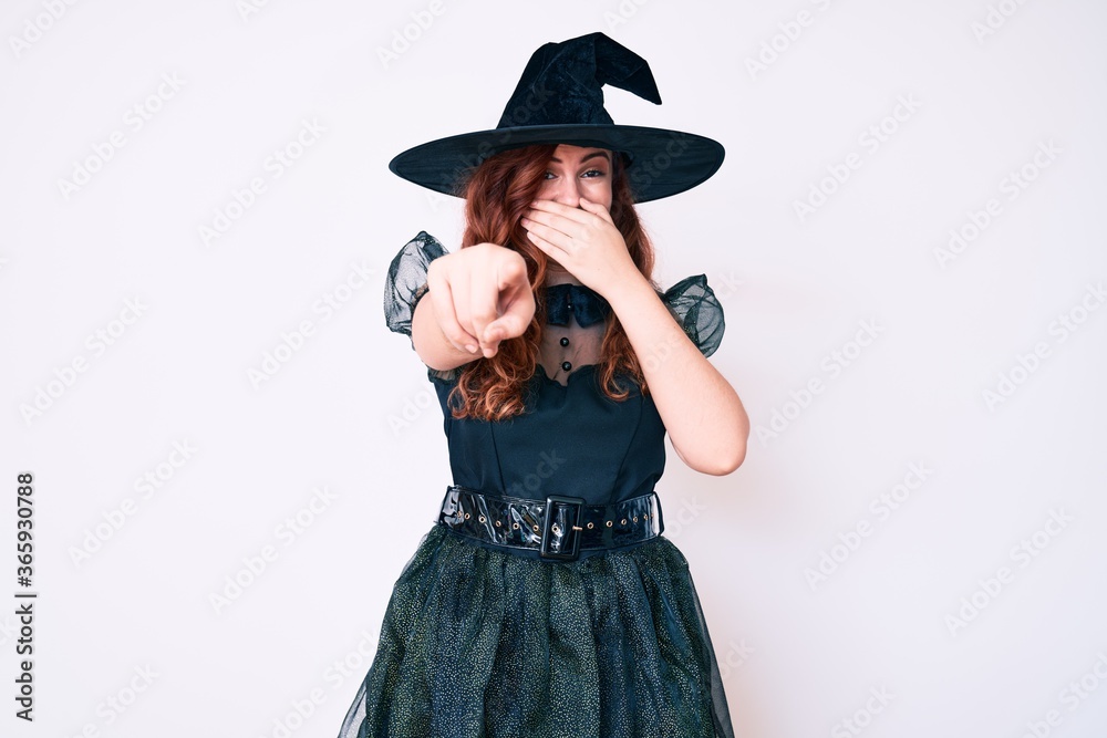 Young beautiful woman wearing witch halloween costume laughing at you, pointing finger to the camera with hand over mouth, shame expression Young beautiful woman wearing witch halloween costume laughing at you, pointing finger to the camera with hand over mouth, shame expression