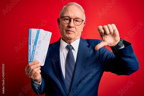 Senior grey haired business man holding airplane boarding pass over red background with angry face, negative sign showing dislike with thumbs down, rejection concept