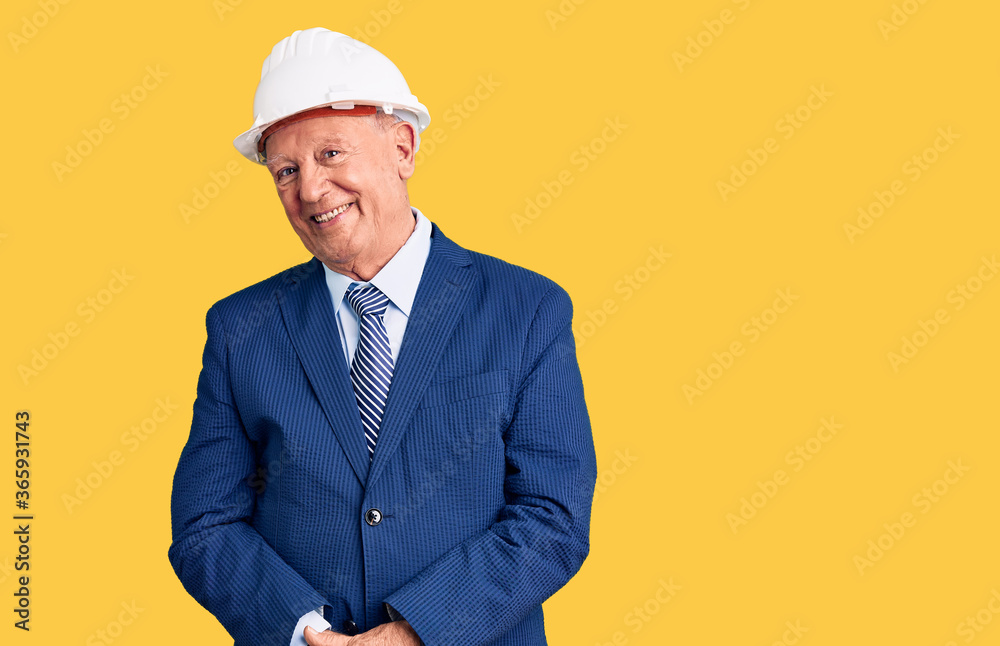 Senior handsome grey-haired man wearing suit and architect hardhat looking away to side with smile on face, natural expression. laughing confident.