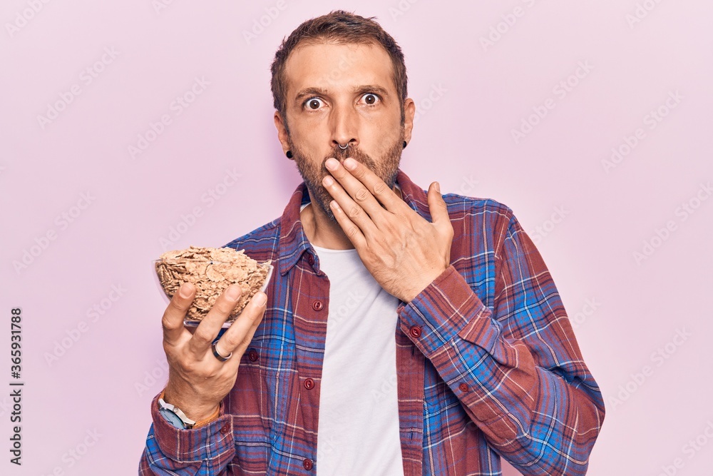 Young handsome man holding cornflakes covering mouth with hand, shocked and afraid for mistake. surprised expression Young handsome man holding cornflakes covering mouth with hand, shocked and afraid for mistake. surprised expression