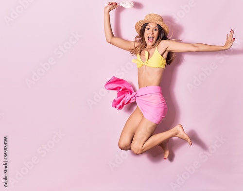 Young beautiful girl wearing bikini and summer hat smiling happy. Jumping with smile on face holding lollipop over isolated pink background