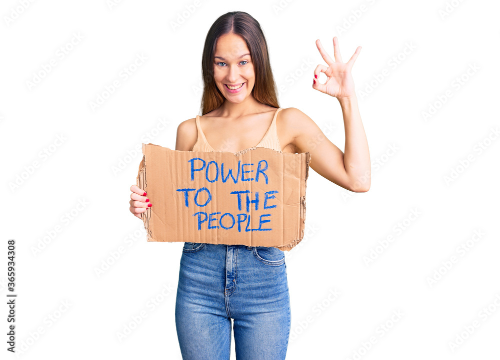 Beautiful brunette young woman holding power to the people banner doing ok sign with fingers, smiling friendly gesturing excellent symbol Beautiful brunette young woman holding power to the people banner doing ok sign with fingers, smiling friendly gesturing excellent symbol