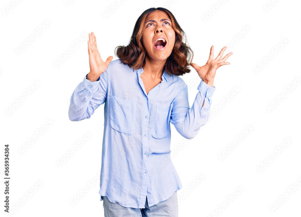 Young beautiful mixed race woman wearing casual business shirt crazy and mad shouting and yelling with aggressive expression and arms raised. frustration concept. Young beautiful mixed race woman wearing casual business shirt crazy and mad shouting and yelling with aggressive expression and arms raised. frustration concept.
