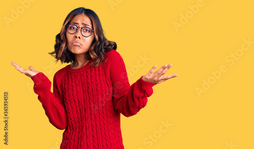 Young beautiful mixed race woman wearing red sweater and glasses clueless and confused expression with arms and hands raised. doubt concept.
