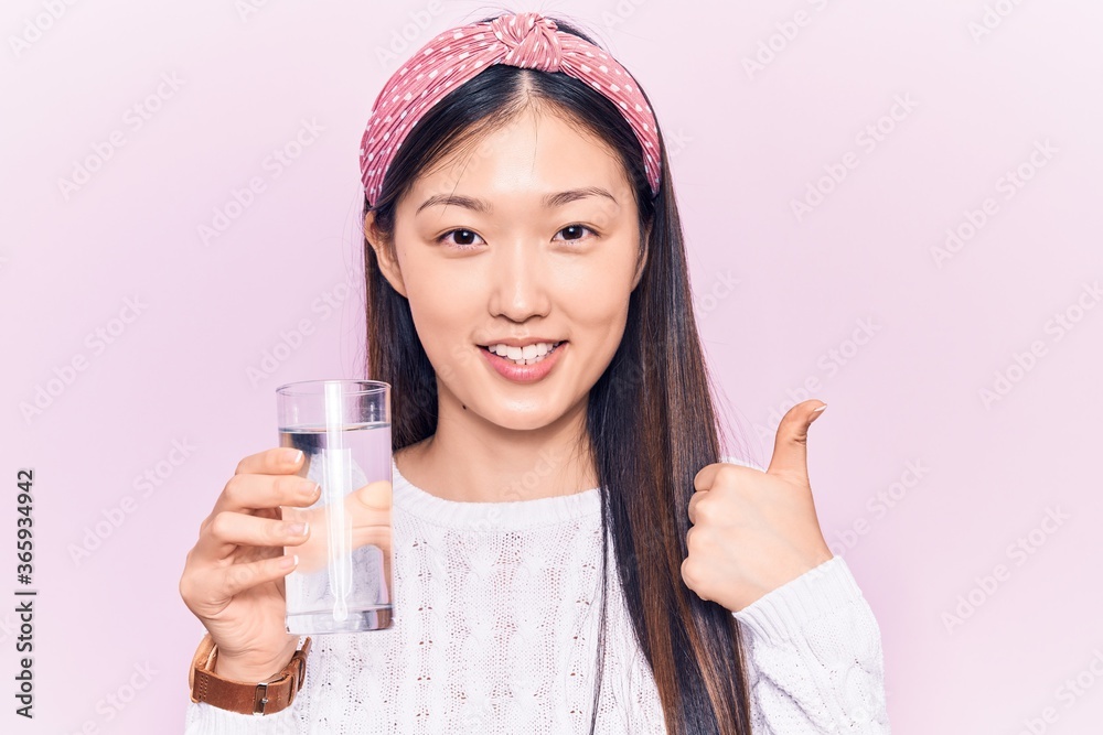 Young beautiful chinese woman drinking glass of water smiling happy and positive, thumb up doing excellent and approval sign Young beautiful chinese woman drinking glass of water smiling happy and positive, thumb up doing excellent and approval sign