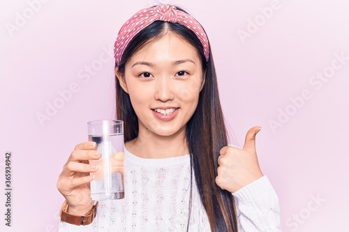 Young beautiful chinese woman drinking glass of water smiling happy and positive, thumb up doing excellent and approval sign