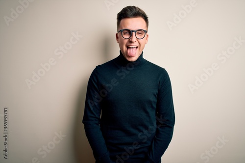 Young handsome caucasian man wearing glasses and casual sweater over isolated background sticking tongue out happy with funny expression. Emotion concept.