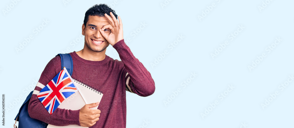 Fototapeta premium Hispanic handsome young man exchange student holding uk flag smiling happy doing ok sign with hand on eye looking through fingers