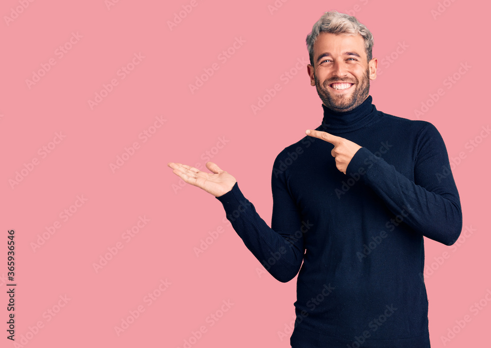 Young handsome blond man wearing casual turtleneck sweater amazed and smiling to the camera while presenting with hand and pointing with finger.