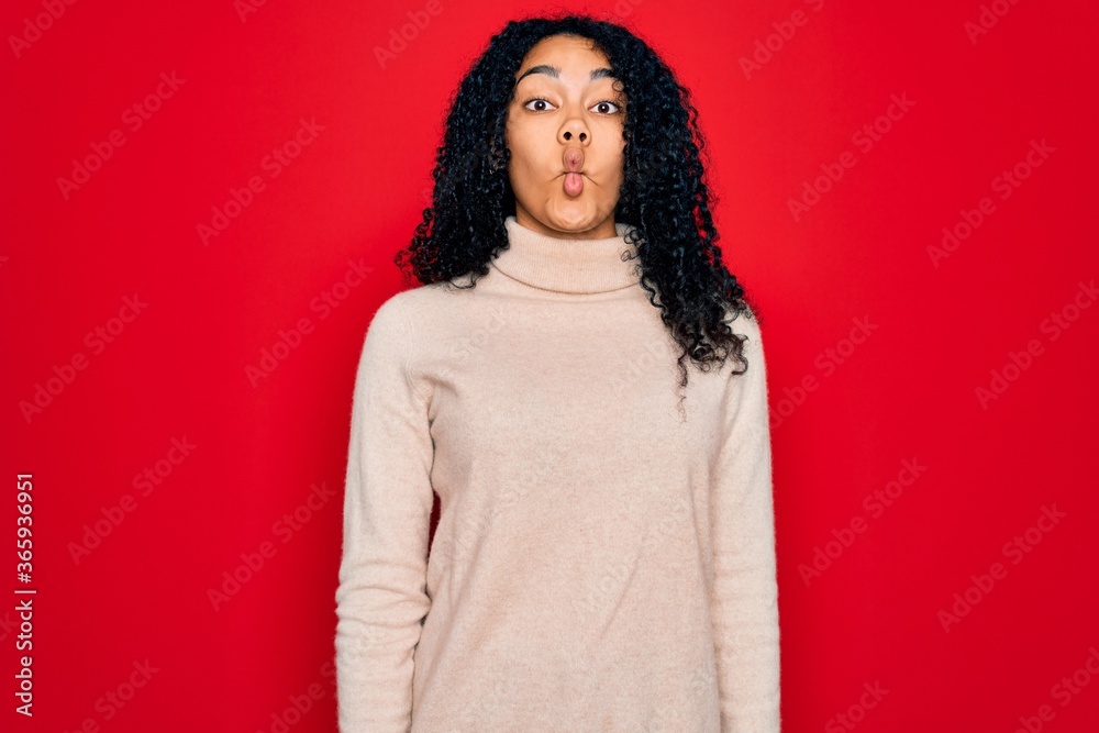 Fototapeta premium Young african american curly woman wearing casual turtleneck sweater over red background making fish face with lips, crazy and comical gesture. Funny expression.