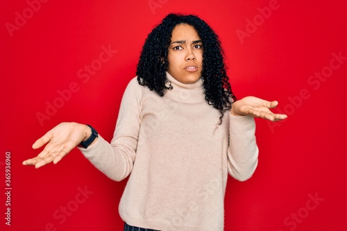 Canvas Print Young african american curly woman wearing casual turtleneck sweater over red background clueless and confused with open arms, no idea concept
