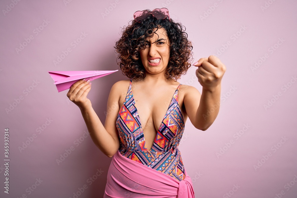 Young beautiful curly arab woman on vacation wearing swimsuit holding pink paper airplane annoyed and frustrated shouting with anger, crazy and yelling with raised hand, anger concept Young beautiful curly arab woman on vacation wearing swimsuit holding pink paper airplane annoyed and frustrated shouting with anger, crazy and yelling with raised hand, anger concept