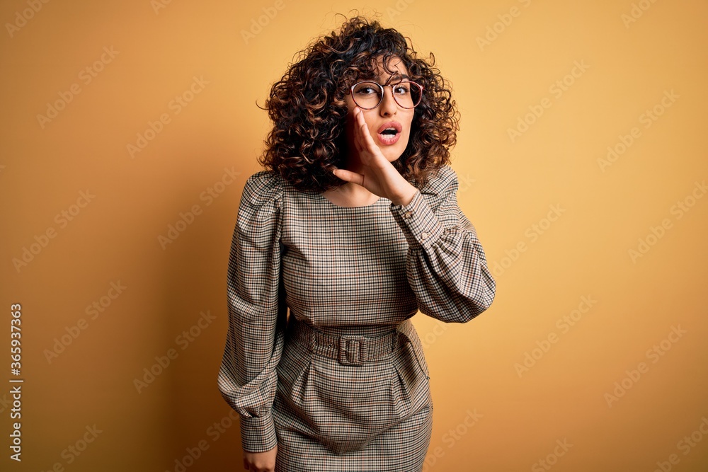 Beautiful arab business woman wearing dress and glasses standing over yellow background hand on mouth telling secret rumor, whispering malicious talk conversation