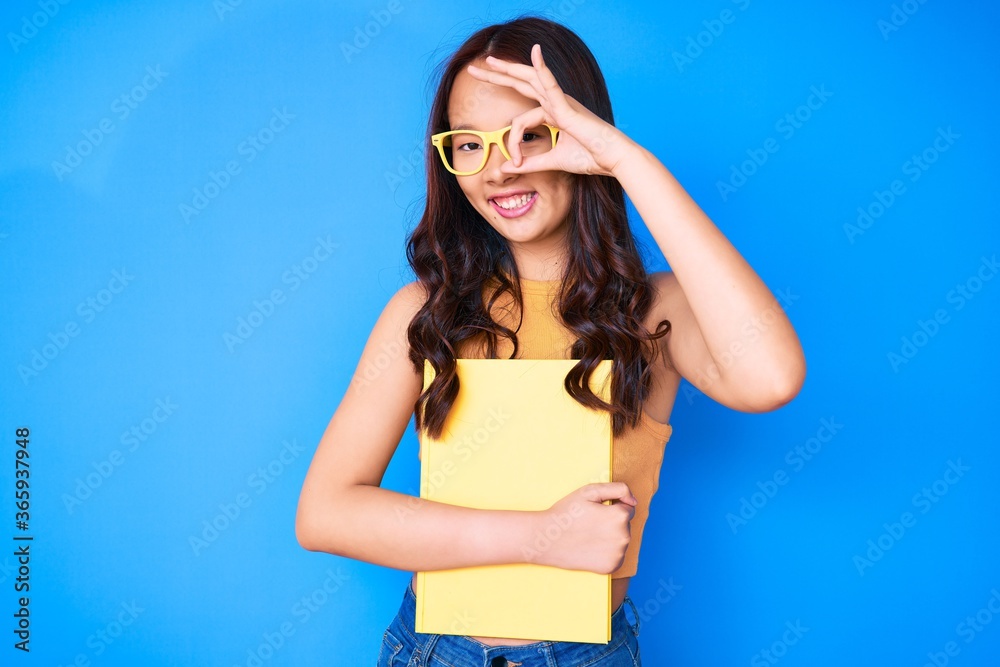 Young beautiful chinese girl wearing glasses holding book smiling happy doing ok sign with hand on eye looking through fingers Young beautiful chinese girl wearing glasses holding book smiling happy doing ok sign with hand on eye looking through fingers