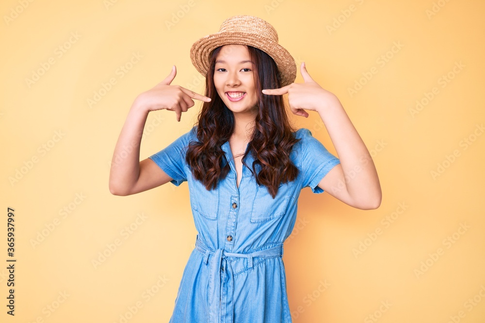 Young beautiful chinese girl wearing summer hat smiling cheerful showing and pointing with fingers teeth and mouth. dental health concept. Young beautiful chinese girl wearing summer hat smiling cheerful showing and pointing with fingers teeth and mouth. dental health concept.