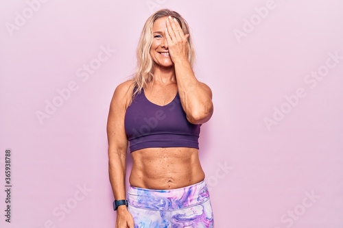 Middle age caucasian blonde woman wearing sportswear over pink background covering one eye with hand  confident smile on face and surprise emotion.
