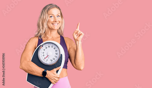 Middle age fit blonde woman wearing sports clothes holding weighing machine surprised with an idea or question pointing finger with happy face, number one