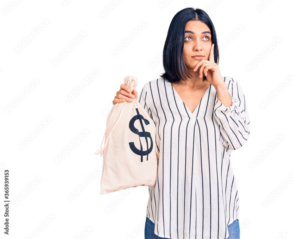 Young beautiful latin woman holding money bag with dollar symbol serious face thinking about question with hand on chin, thoughtful about confusing idea